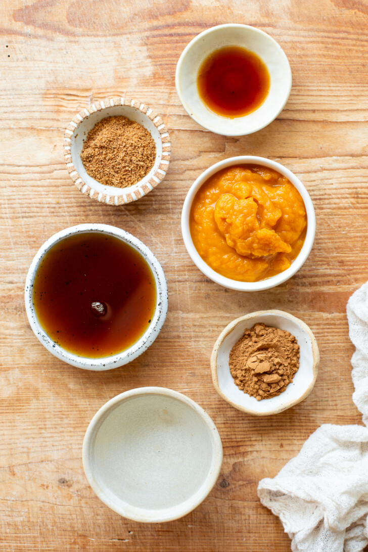 Ingredients for pumpkin spice syrup in bowls: coconut sugar, vanilla extract, maple syrup, pumpkin puree, water, and pumpkin pie spice on a wooden bread board.